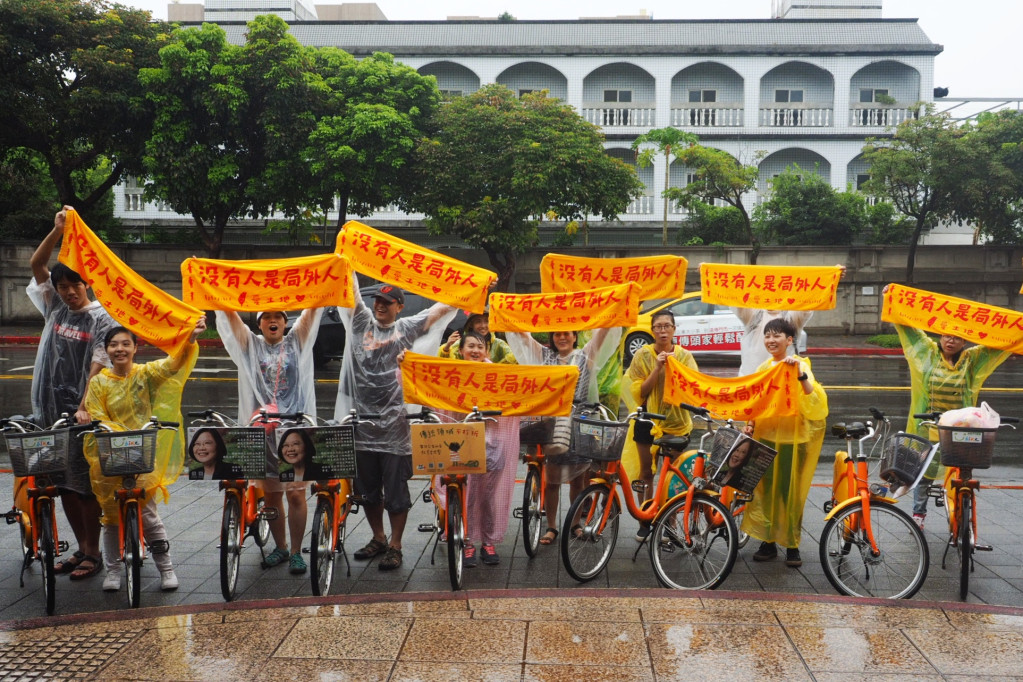 民眾冒著風雨騎腳踏車，將日前被警方清運的石頭從公館寶藏巖送回凱道旁的台大醫院站出口，高舉「沒有人是局外人」的毛巾合照。圖／張家銘